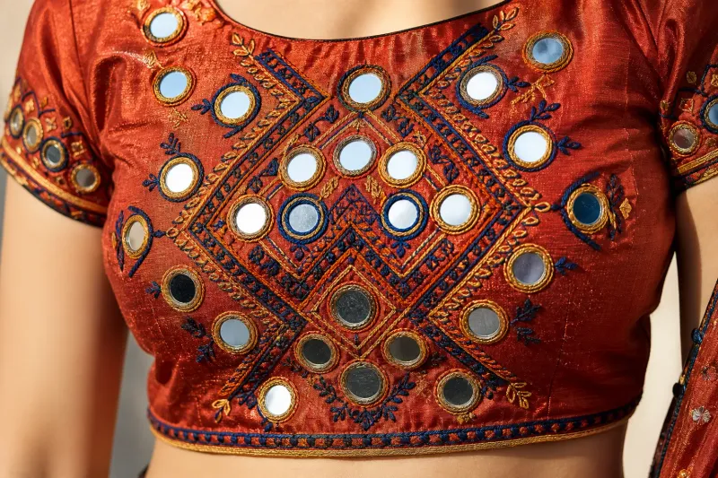 kutch-mirror-work-107fa86d Macro close-up of a designer blouse with Kutch mirror-work — rust, navy, and gold geometric silk embroidery around small circular mirrors on textured raw silk.
