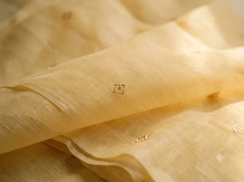 macro-close-up-ca23561a Macro close-up of pastel marigold yellow Chanderi silk-cotton fabric with fine gold zari bootis, catching soft natural sunlight.